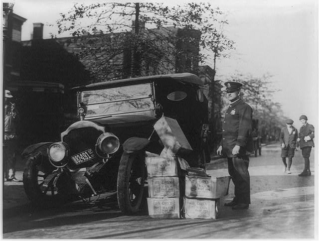 Policeman standing alongside wrecked car and cases of moonshine